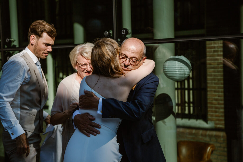 Knuffel bruid en vader tijdens het diner in het Muntgebouw Utrecht met avondlicht tussen de ramen.
